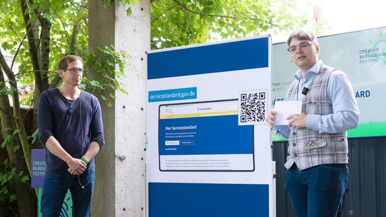 Unter Bäumen stehen zwei junge Männer mit einer großen Präsentationstafel, auf der ein Screenshot der neuen Service-Standard-Website gezeigt wird.