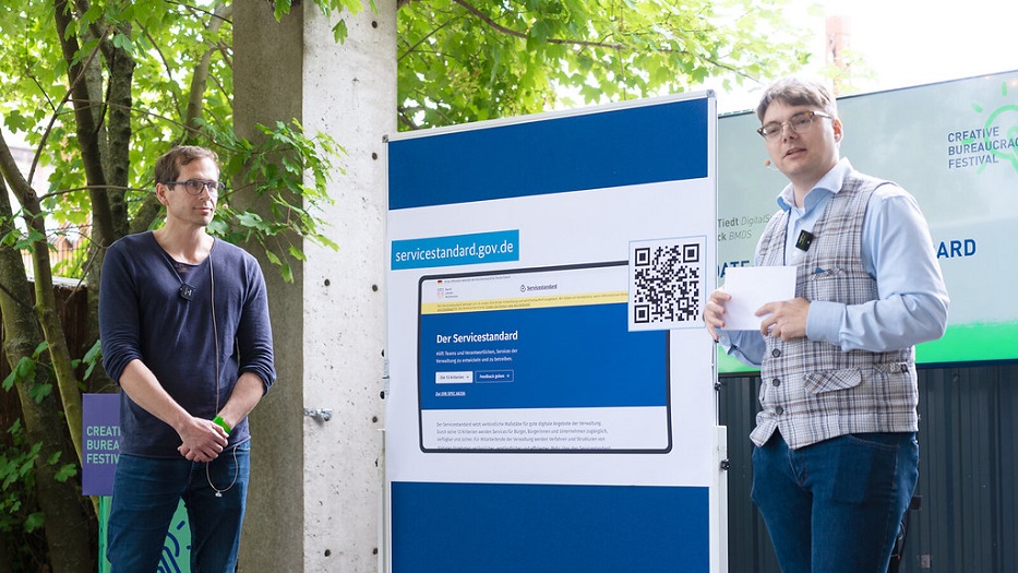 Unter Bäumen stehen zwei junge Männer mit einer großen Präsentationstafel, auf der ein Screenshot der neuen Service-Standard-Website gezeigt wird.