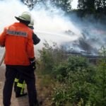 Feuerwehrmann mit einen Schlauch in der Hand steht vor brennendem Gras an einer Böschung.