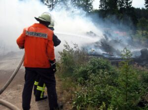 Feuerwehrmann mit einen Schlauch in der Hand steht vor brennendem Gras an einer Böschung.
