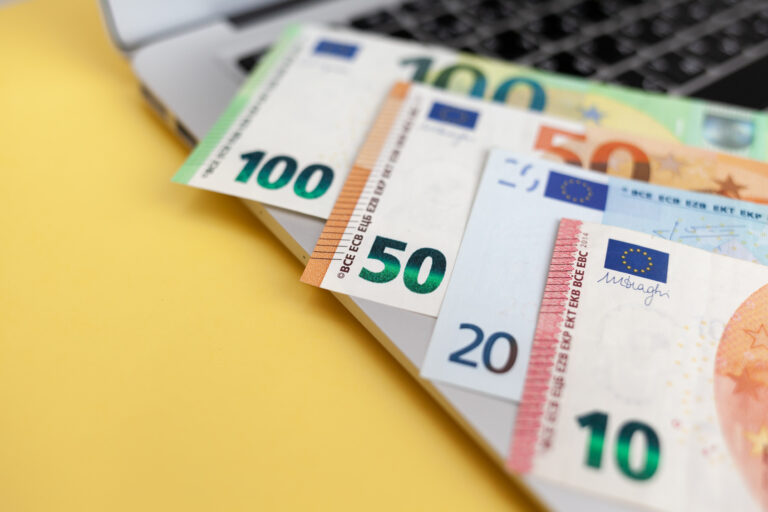 Notebook auf gelber Tischplatte, darauf liegen Bündel von Euro-Scheinen in variierter Stückelung.