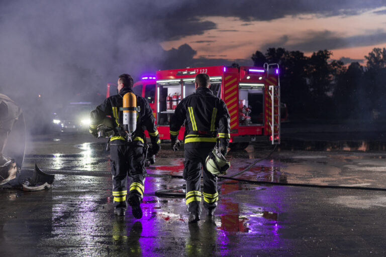 Mutmaßlicher Feuerehreinsatz im Abendlicht, man sieht zwei Einsatzkräfte von hinten durch eine Pfütze auf ihr Fahrzeug zulaufen, im Hintergrund Rauch und Lichter.