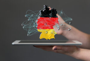 Composit: Hände halten ein Tablet, darüber schwebt ein 2D-Objekt mit den Umrissen und Farben von Deutschland, überlagert von einem gezeichneten Netzwerk.