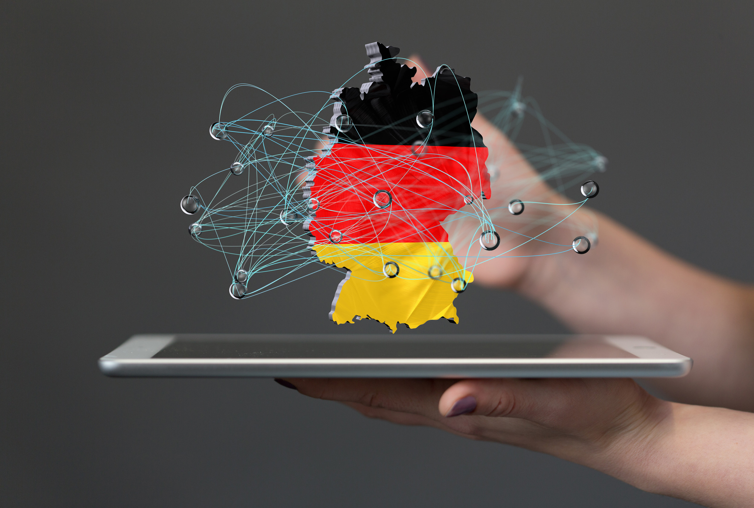 Composit: Hände halten ein Tablet, darüber schwebt ein 2D-Objekt mit den Umrissen und Farben von Deutschland, überlagert von einem gezeichneten Netzwerk.