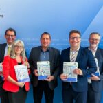 Gruppenfoto von förmlich gekleideten Männern und einer Frau vor einer blauen Wand.