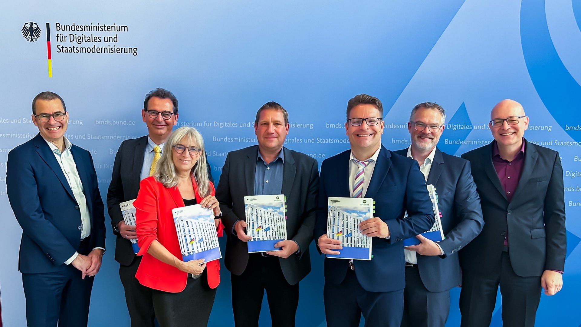 Gruppenfoto von förmlich gekleideten Männern und einer Frau vor einer blauen Wand.