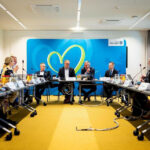 Große, nüchterner Raum mit U-förmiger Tischanordnung, an der Menschen in Anzügen sitzen.