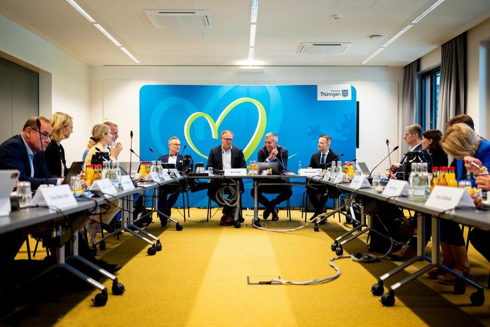 Große, nüchterner Raum mit U-förmiger Tischanordnung, an der Menschen in Anzügen sitzen.