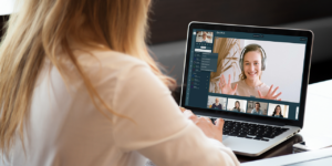 Blick über die Schulter einer jungen Frau, auf deren Laptop eine Videokonferenz läuft.