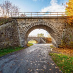 Schmale Asphaltstraße, die unter einer engen, gemauerten Rundbogen-Brücke hindurchführt.