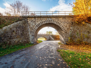 Schmale Asphaltstraße, die unter einer engen, gemauerten Rundbogen-Brücke hindurchführt.