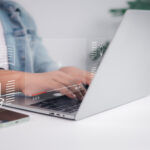 Oberkörper und Häne einer Person in einer Büroumgebung, die auf einem silbernen Laptop tippt.