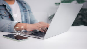 Oberkörper und Häne einer Person in einer Büroumgebung, die auf einem silbernen Laptop tippt.