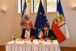 Zwei Männer in dunklen Anzügen sitzen vor Fahnen an einem Tisch und unterzeichnen Papiere.