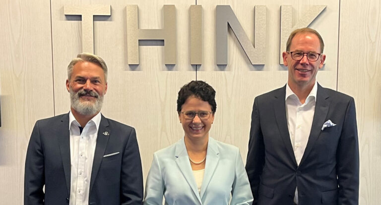 Zwei Männer und eine Frau in formaler Kleidung stehen lächelnd aufgereiht vor einer Wand mit dem Wort "Think".