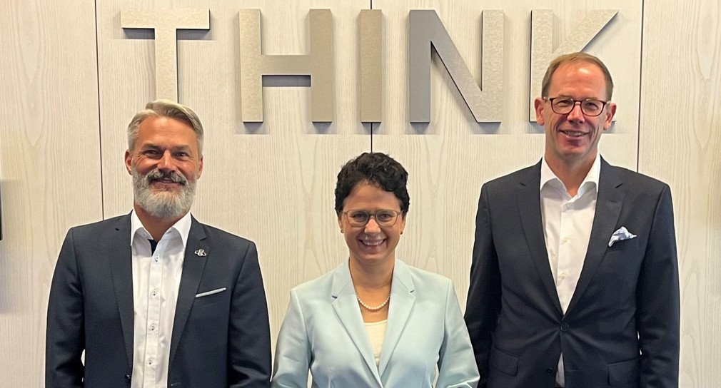 Zwei Männer und eine Frau in formaler Kleidung stehen lächelnd aufgereiht vor einer Wand mit dem Wort "Think".