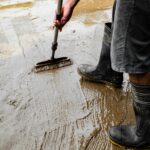 Durch Hochwasser verschlammter Fußboden in einem Haus, eine Person mit Gummistiefeln macht sauber.