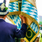 Rückansicht eines Mannes in bayerischer Tracht, der eine Fahne mit blau-weißem Rautenmuster festhält.