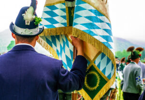 Rückansicht eines Mannes in bayerischer Tracht, der eine Fahne mit blau-weißem Rautenmuster festhält.