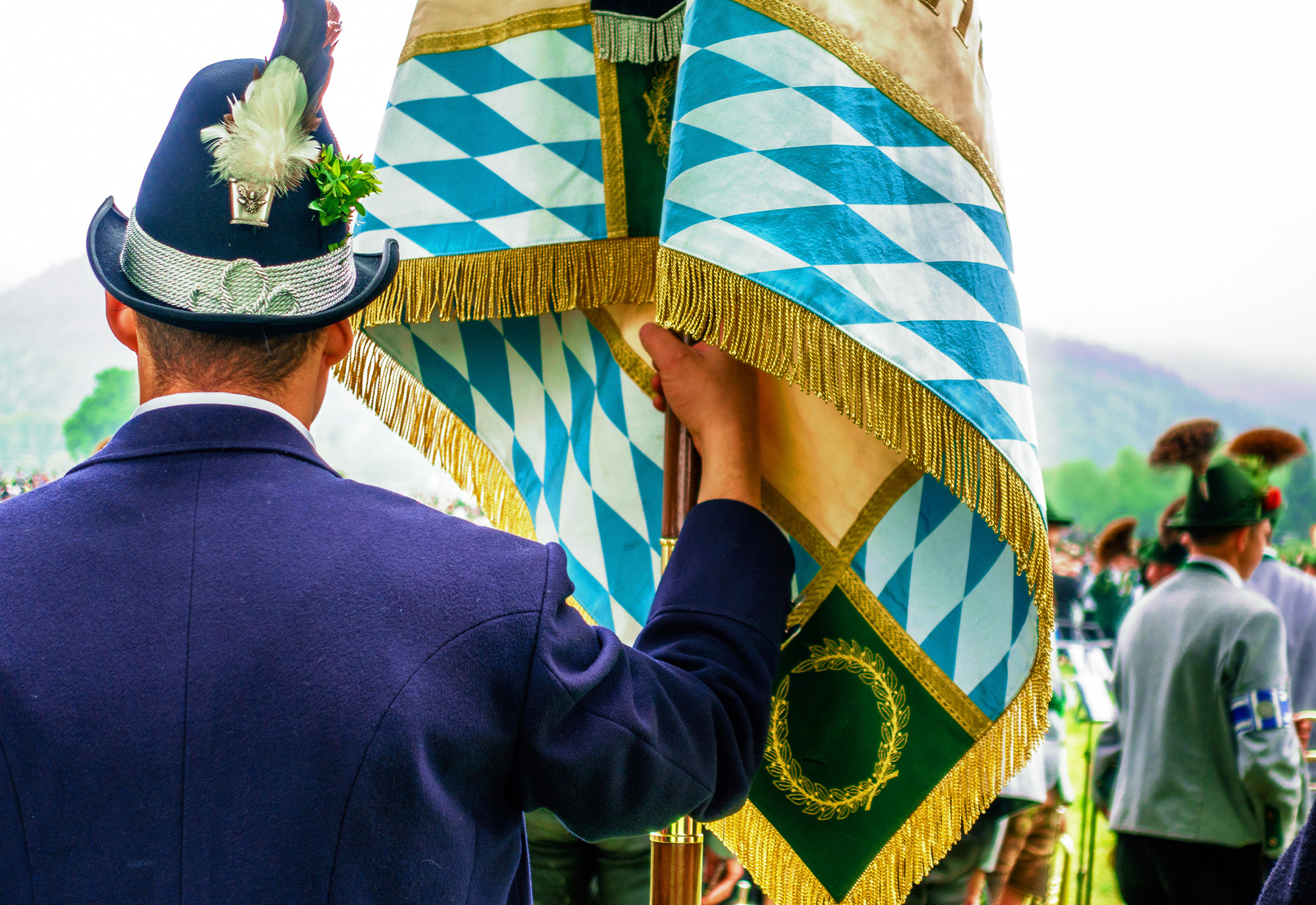 Rückansicht eines Mannes in bayerischer Tracht, der eine Fahne mit blau-weißem Rautenmuster festhält.