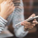 Hände und Torso einer mutmaßlich weiblichen Person, die mit nachdenklicher Pose ein Smartphone hält.