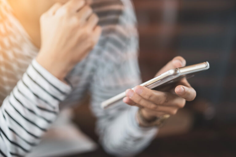 Hände und Torso einer mutmaßlich weiblichen Person, die mit nachdenklicher Pose ein Smartphone hält.