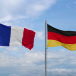 Französische und deutsche Flagge wehen nebeneinander, im Hintergrund leicht bewölkter blauer Himmel.