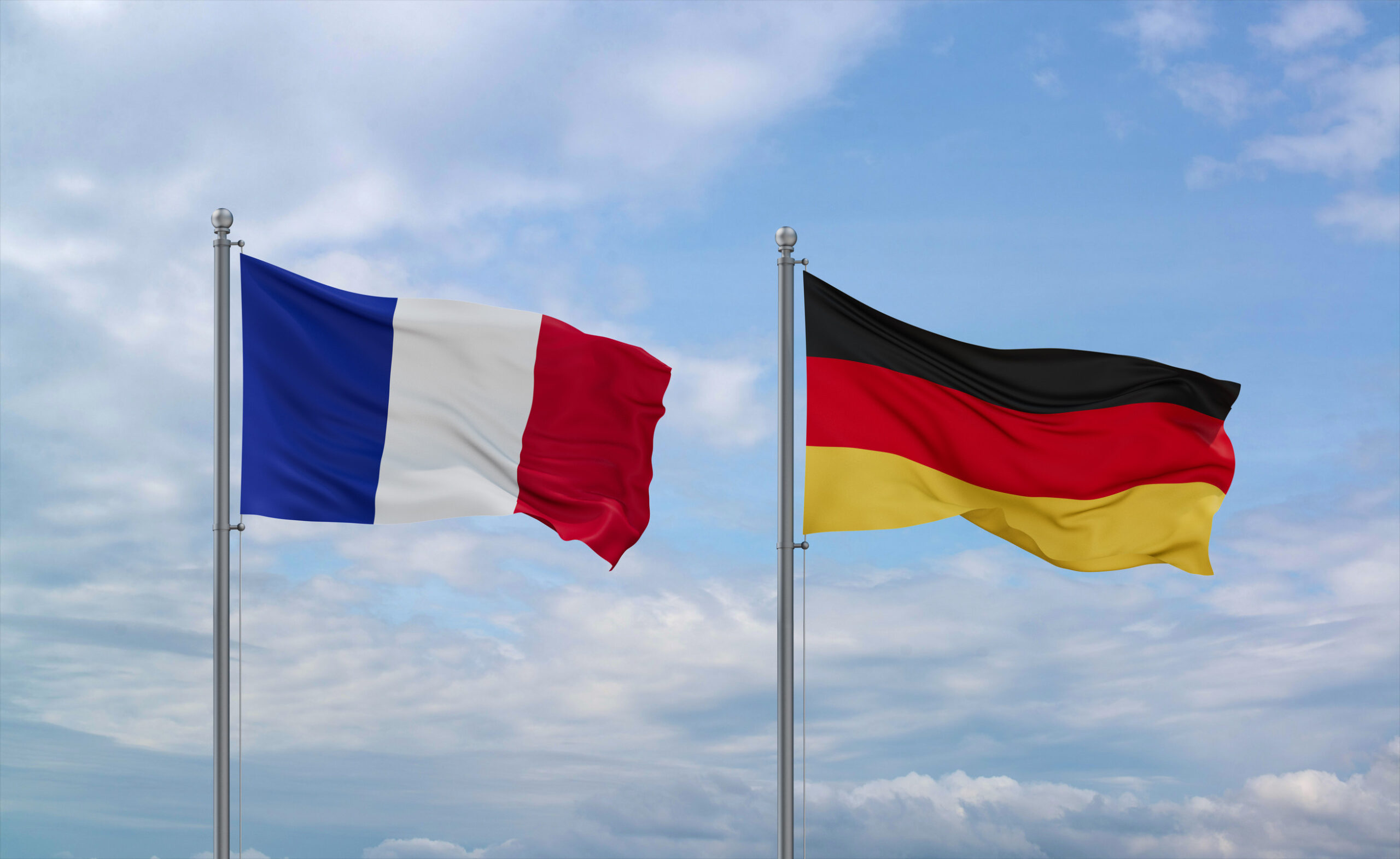 Französische und deutsche Flagge wehen nebeneinander, im Hintergrund leicht bewölkter blauer Himmel.