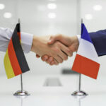 Zwei männliche Hände und Arme kommen über einem Konferenztisch zu einem Handshake zusammen, im Vordergrund Minitaur-Nationalflagen von Deutschland und Frankreich.