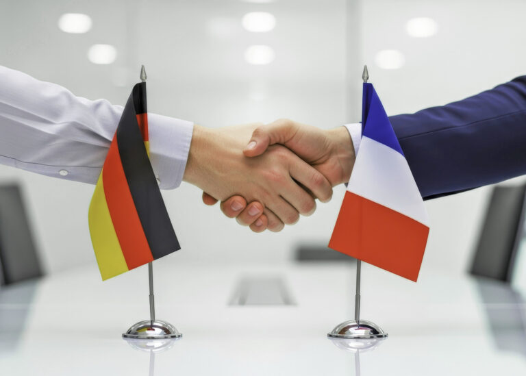 Zwei männliche Hände und Arme kommen über einem Konferenztisch zu einem Handshake zusammen, im Vordergrund Minitaur-Nationalflagen von Deutschland und Frankreich.