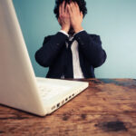 Ein Schreibtisch mit aufgeklapptem Laptop, dahinter sitzt ein mann, der sich die Hände vors Gesicht schlägt.