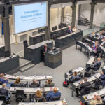 Parlamentarischer Sitzungssaal mit besetzten Sitzreihen in Hufeisenform, am Podium steht ein Redner.
