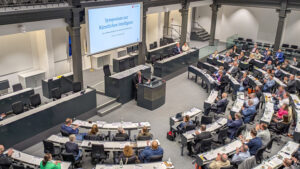 Parlamentarischer Sitzungssaal mit besetzten Sitzreihen in Hufeisenform, am Podium steht ein Redner.