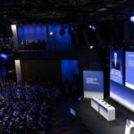 Blick von oben in einen abgedunkelten, großen, hohen Konferenzraum mit viel Publikum. Au der blau beleuchteten Bühne sieht man Karsten Wildberger.