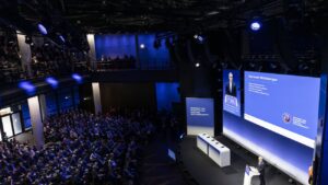 Blick von oben in einen abgedunkelten, großen, hohen Konferenzraum mit viel Publikum. Au der blau beleuchteten Bühne sieht man Karsten Wildberger.