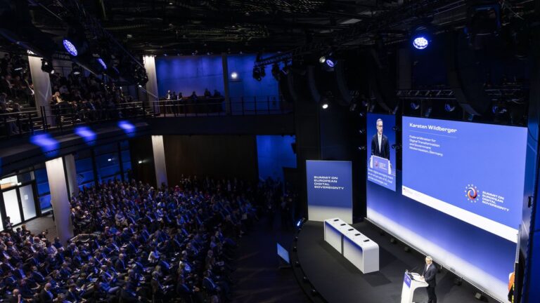 Blick von oben in einen abgedunkelten, großen, hohen Konferenzraum mit viel Publikum. Au der blau beleuchteten Bühne sieht man Karsten Wildberger.