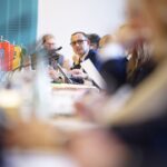 Blick über vollbesetzten Konferenztisch mit Landeswimpeln, großer Unschärfebereich im Vordergrund, Fokus iegt auf Schleswig-Holsteins Digitalminister Dirk Schrödter