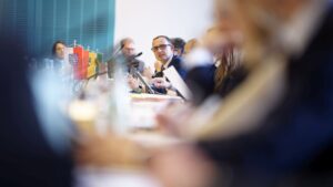 Blick über vollbesetzten Konferenztisch mit Landeswimpeln, großer Unschärfebereich im Vordergrund, Fokus iegt auf Schleswig-Holsteins Digitalminister Dirk Schrödter