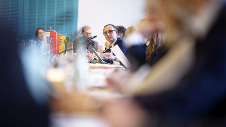 Blick über vollbesetzten Konferenztisch mit Landeswimpeln, großer Unschärfebereich im Vordergrund, Fokus iegt auf Schleswig-Holsteins Digitalminister Dirk Schrödter