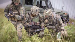 Drei Soldaten hocken auf einer Wiese. Ein Soldat bedient ein Tablet. Im Hintergrund steht ein Fahrzeug.