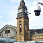 Blick auf ein Bahnhofsgebäude, im Vordergrund ein Laternenmast mit montierter Kamera.