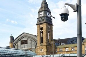 Blick auf ein Bahnhofsgebäude, im Vordergrund ein Laternenmast mit montierter Kamera.