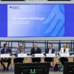 Podium mit sieben Personen an einem langen Tisch, davor die Schultern von Publikum, dahinter eine "BMDS"-Projektion.