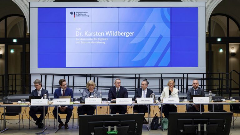 Podium mit sieben Personen an einem langen Tisch, davor die Schultern von Publikum, dahinter eine "BMDS"-Projektion.