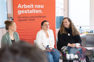 Drei Frauen sitzen vor einem Plakat mit der Aufschrift "Arbeit neu gestalten"