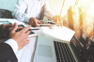 Detailansicht eines moernen Schreibtischs mit Laptop, man sieht zwei Männertorsos, die etwas zusammen bearbeiten.