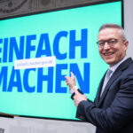 Karsten Wildberger mit sichtlich guter Laune vor einem großen Screen auf dem "EINFACH MACHEN" steht.