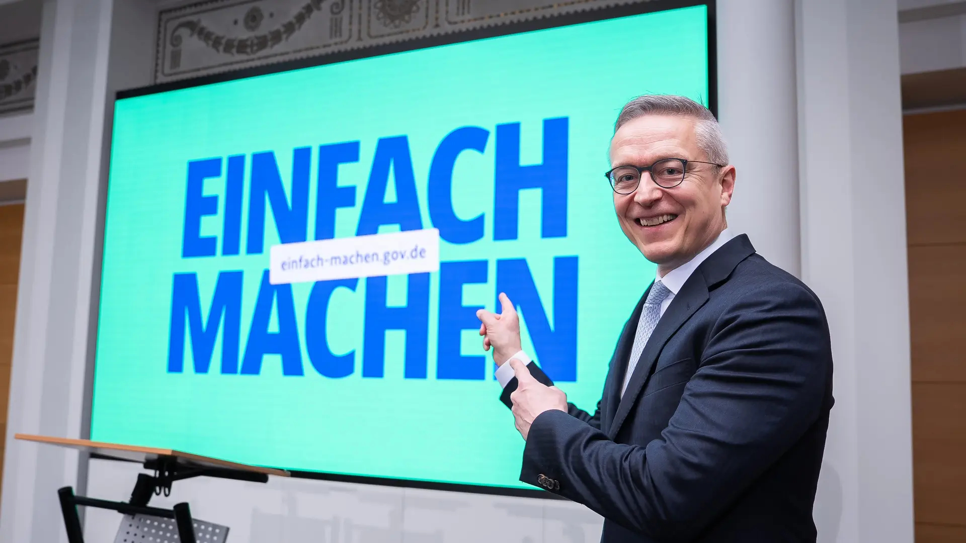 Karsten Wildberger mit sichtlich guter Laune vor einem großen Screen auf dem "EINFACH MACHEN" steht.