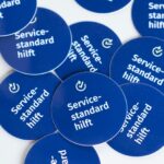 Größere Menge von blauen, runden Aufklebern mit der Aufschrift "Servicestandard hilft", die auf einer weißen Tischplatte liegen.