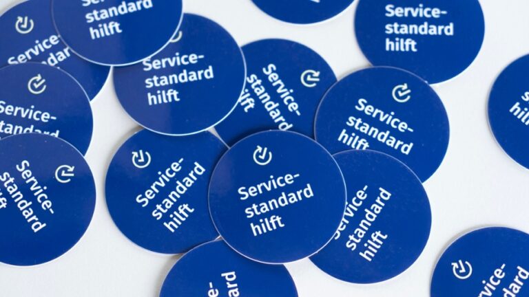 Größere Menge von blauen, runden Aufklebern mit der Aufschrift "Servicestandard hilft", die auf einer weißen Tischplatte liegen.